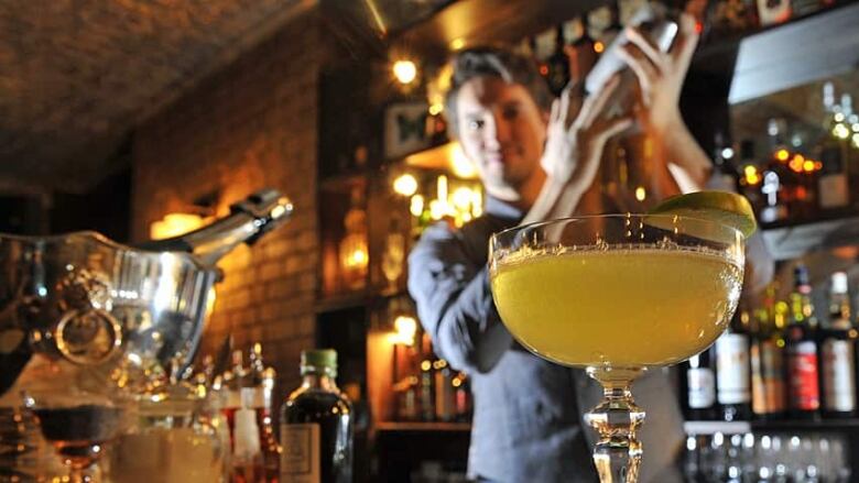 A margarita sits on a bar in the foreground, while behind it, a bartender uses a cocktail shaker. In the background are bottles on a shelf and other out-of-focus bar accoutrements.