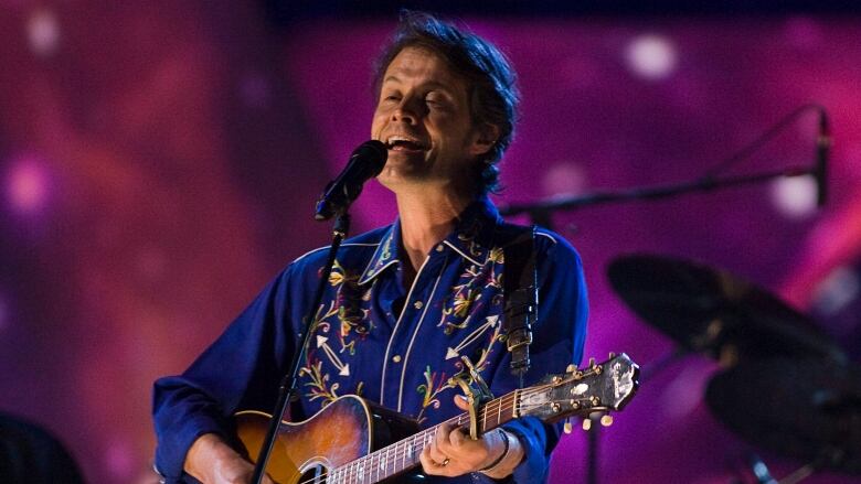 A man in a blue shirt strums a guitar while singing into a microphone. 