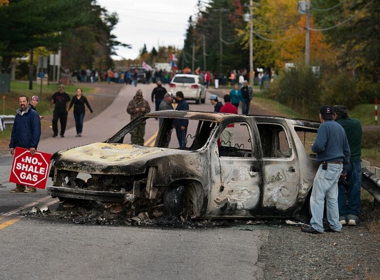 Protesters stand near a burned out vehicle lying on a street.