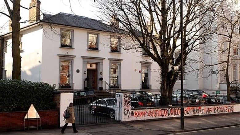 A white building is seen behind a graffitied fence. A large tree stands in front of the building as a pedestrian walks by.