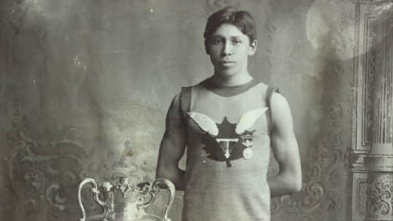 In a black-and-white photo, Tom Longboat stands next to a trophy.