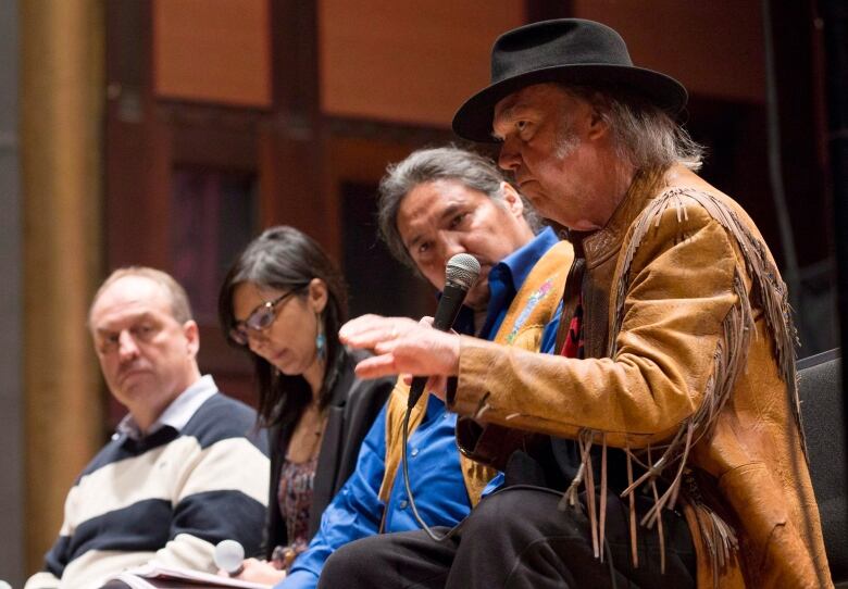 A man in a black hat and buckskin jacket speaks with a microphone while three other people watch.