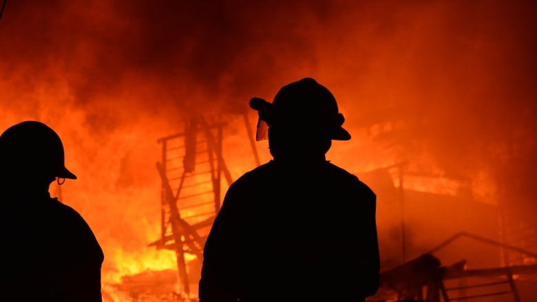 Two firefighters are standing in front of a fire with orange flames in front of a burning building at night.