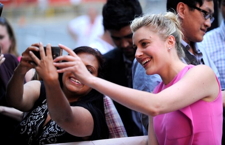 A blond woman wearing pink smiles while taking a photo with a fan.