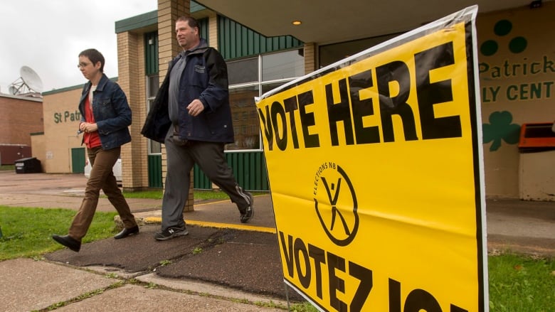 Two people walking out of a polling station, beside a yellow Vote Here sign