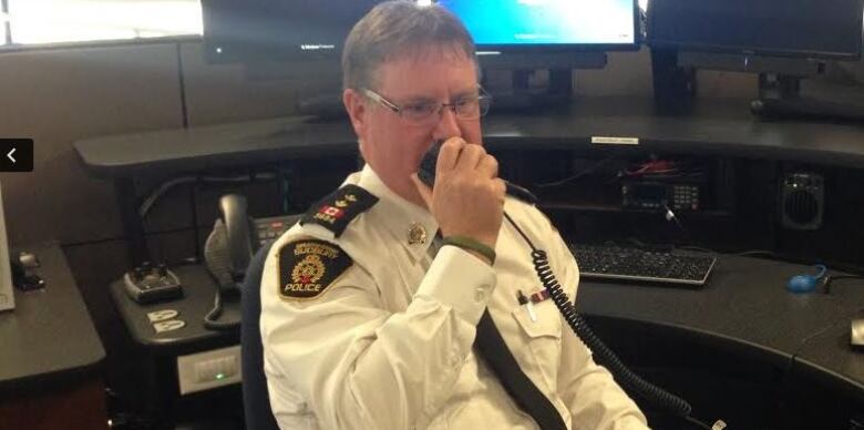 Todd Zimmerman speaks into a police radio, sitting at a desk with several computer screens, wearing a white police uniform