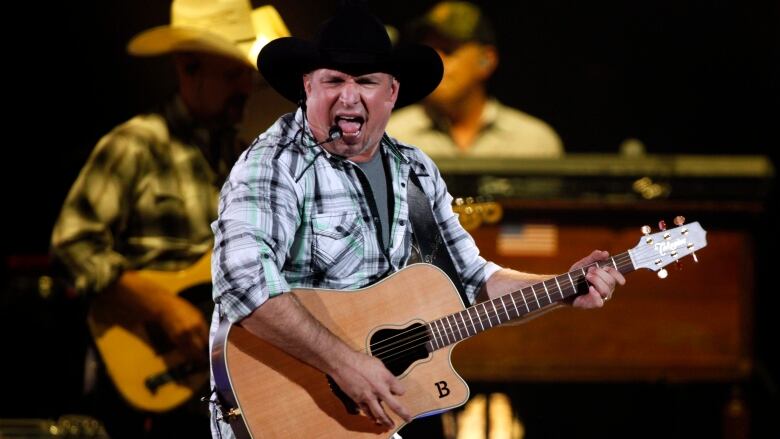 A musician in a black cowboy hat and plaid shirt sings while holding a guitar.