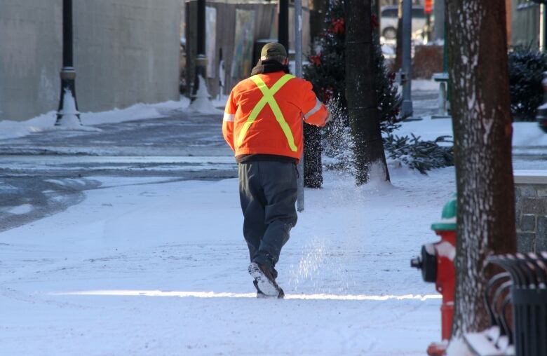 A person in an orange coat throws salt onto a snowy sidewalk