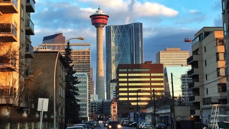 A view of a Calgary Beltline street with cars with their headlights on. Blue sky peaks through clouds. The Calgary Tower looms in the background.