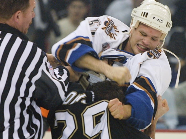 Two hockey players, one wearing white, the other wearing black, fight on the ice as a referee tries to intervene.