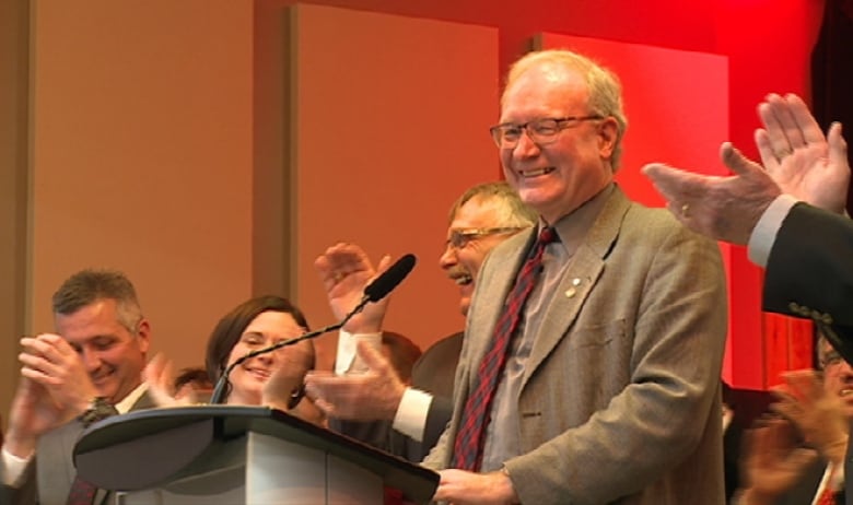 Wade MacLauchlan at podium with people around him applauding.