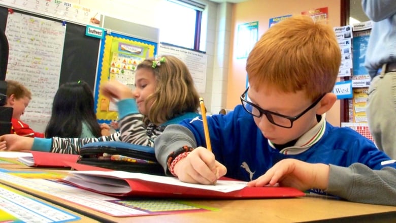 A red-headed student with a pencil writes at his school desk with other students in a classroom with notices and papers pinned to bulletin boards lining the walls.