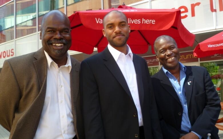 Three men wearing suits are smiling at the camera. Behind them are picnic tables with red umbrellas.