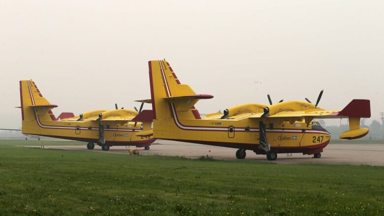 Two yellow-and-red airplanes sit on a tarmac, in front of a large field of grass. The air appears hazy from wildfire smoke.