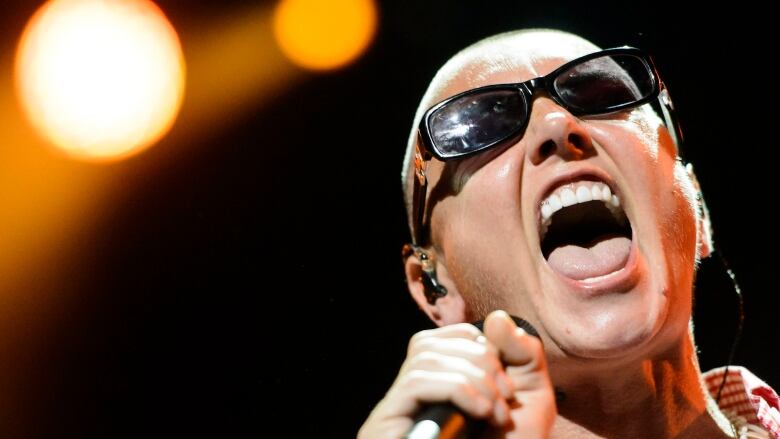 Wearing sunglasses, a woman sings in front of a mic.