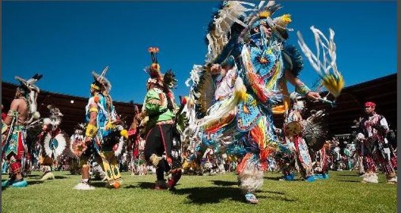 A number of people in Indigenous regalia dance on a grassy area.