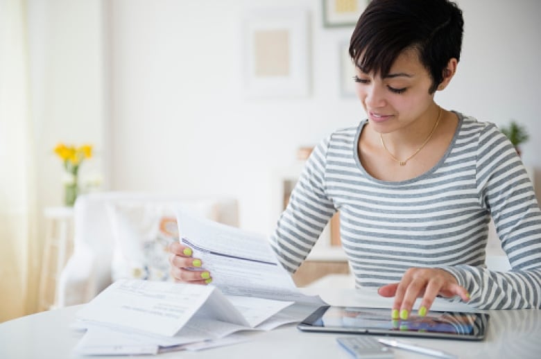 A person with short hair sits at a table and looks over bills while touching a computer tablet.