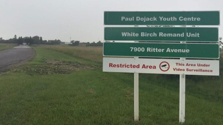 A large green and white sign is shown on a patch of grass, next to a road. It reads: Paul Dojak Youth Centre. White Birch Remand Unit. 7900 Ritter Avenue. Restricted Area: This area under video surveillance.