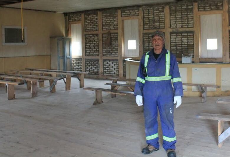 A man in blue coveralls stands in the middle of a brick church with wood pews around him. 