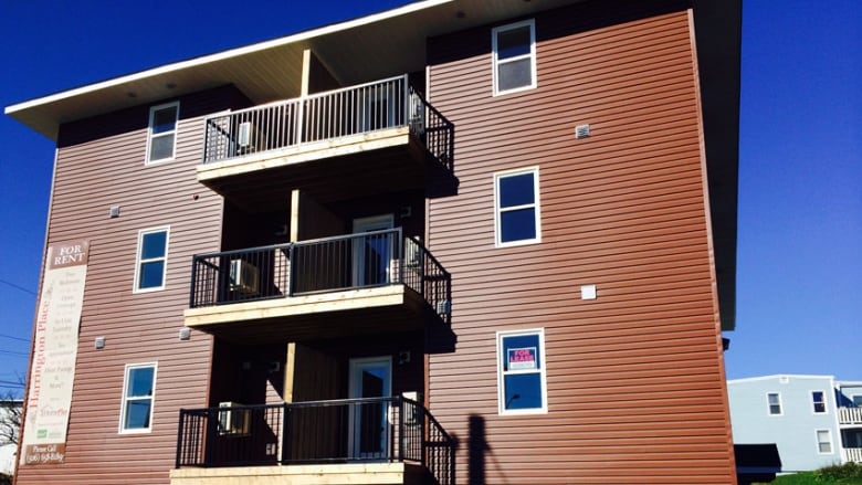 A small three-story apartment building with brown vinyl siding and a balcony in the middle of each floor with a window on each side against a blue sky in the background.