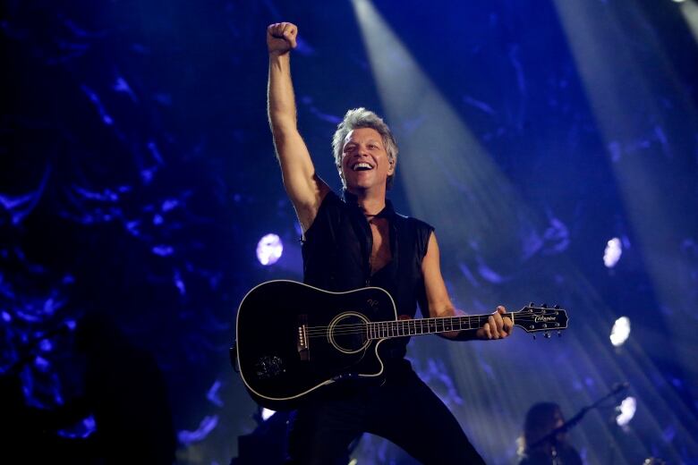 A man in black vest holds a guitar and pumps his fist into the air.