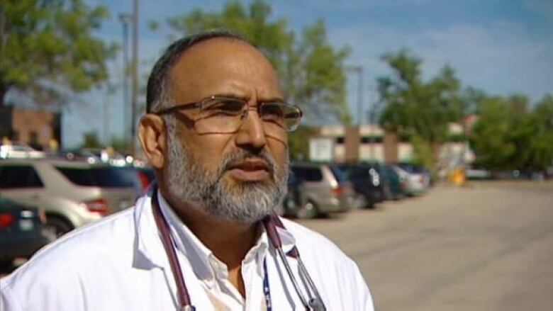 Man with stethoscope around neck stands in parking lot.