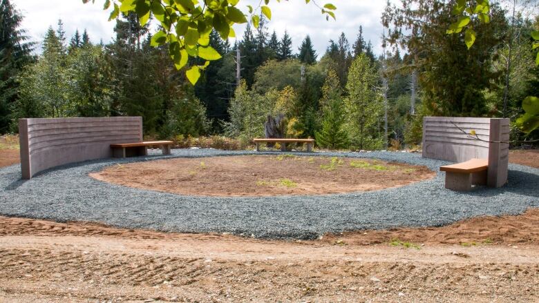 A circle of brown earth with two small benches along perimeter and trees in background in daylight. 