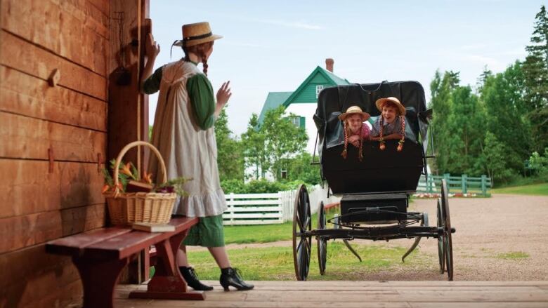 An actor dressed as Anne of Green Gables stands in a barn waving to two girls in Anne hats in an old carriage with the house in the background.