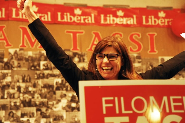 A person with bangs and glasses raises their arms to the sky and smiles with teeth.