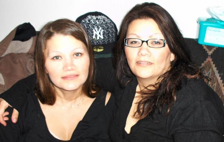 Two women, both with dark hair and dark shirts, sit on a couch together and look into the camera.