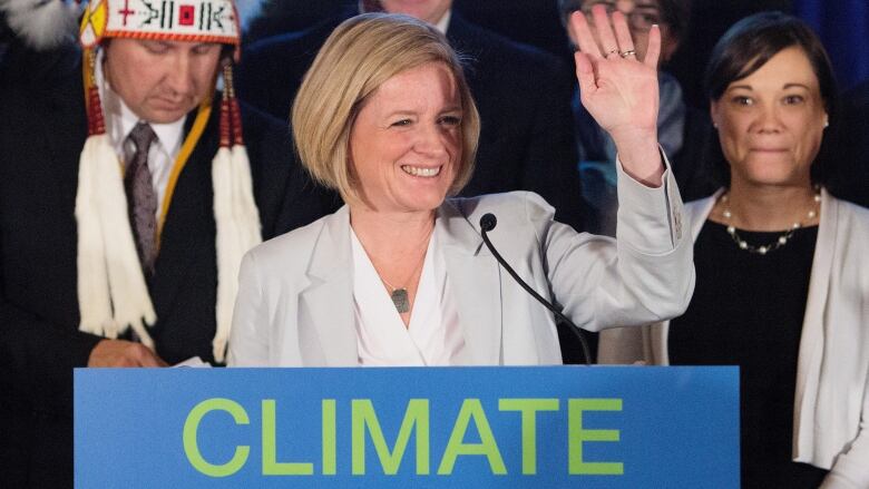 a woman waves behind a lectern. Other people stand around her.