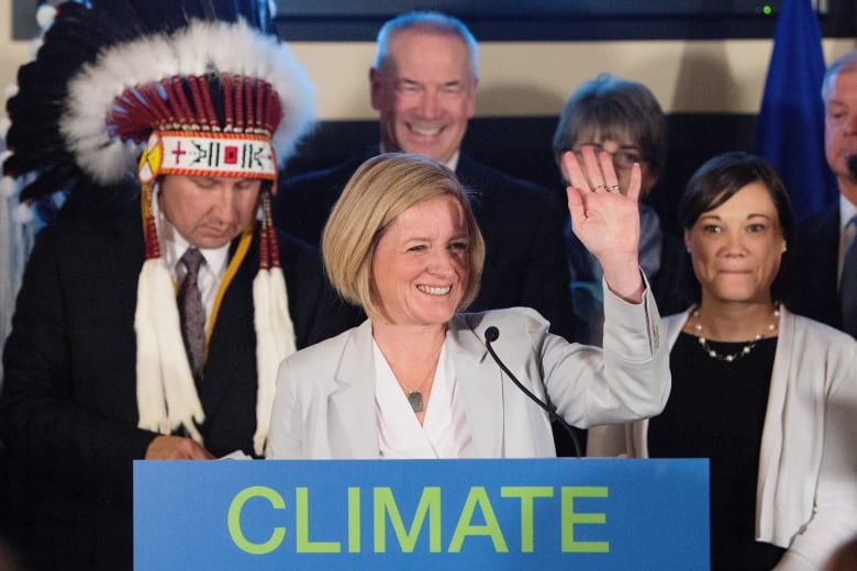 a woman waves behind a lectern. Other people stand around her.