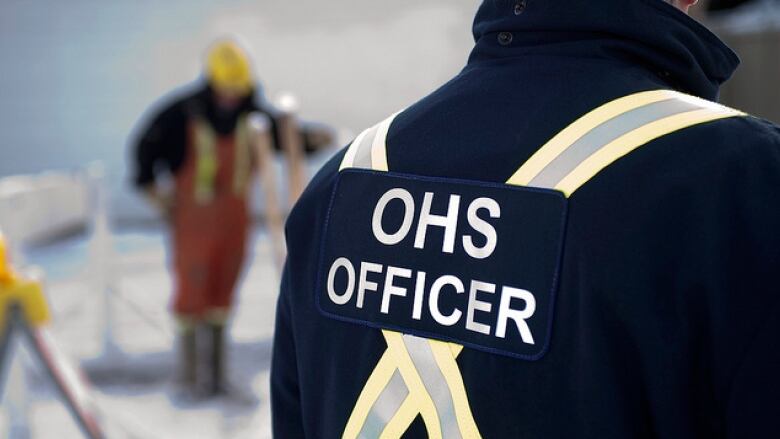 A man stands wearing a blue jacket with a yellow X and the words OHS officer on the back of the jacket.