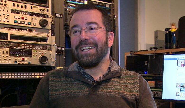 A white man with short brown hair is seen sitting in front of radio equipment in a recording studio