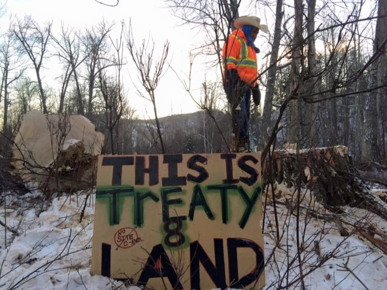 A man wearing a high-vis vest stands on a sign reading 'This is Treaty Land' on a snowy forest.