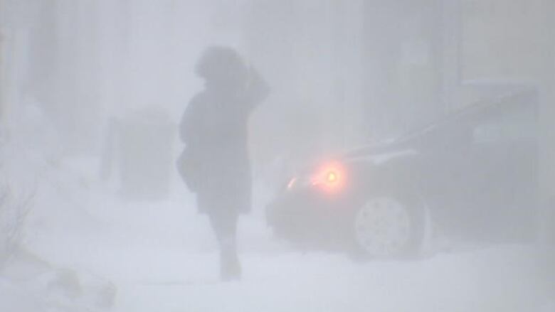 Silhouette of person in parking seen through blowing snow, with vague image of car in the background.