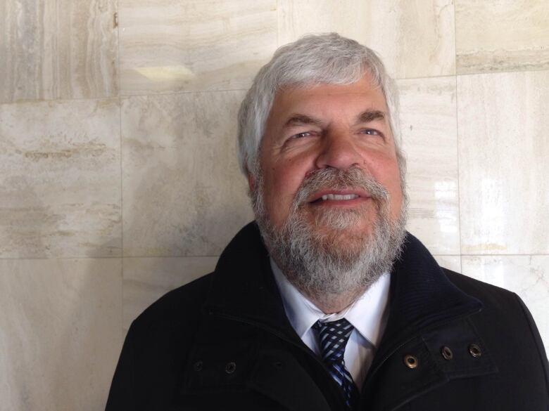 A man with grey hair and beard wearing a black jacket, white shirt and blue tie stands in front of a marble wall. 