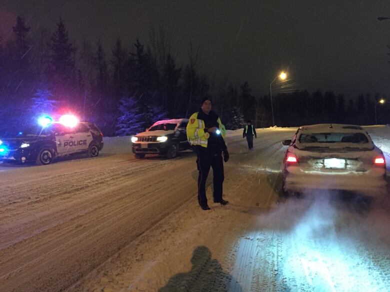 A police officer stands outside in the snow at night, holding a flash light next to a car that is stopped at a RIDE checkpoint.