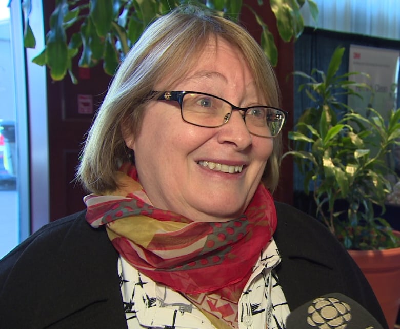 A woman with shoulder-length, dirty-blond hair and glasses is wearing a white blouse, black cardigan and red scarf.