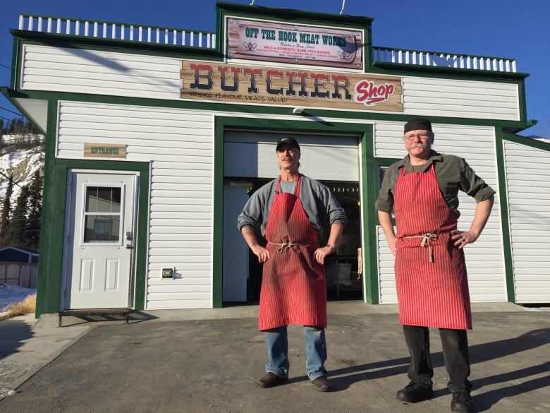 Two men with aprons stand in front of a small one-storey white-and-green building.