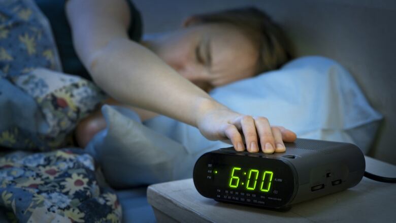 A woman lying in bed with her eyes closed hits the snooze button on an alarm clock reading 6:00. 