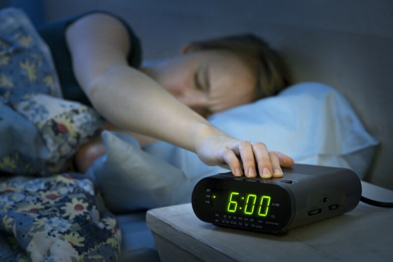 A woman lying in bed with her eyes closed hits the snooze button on an alarm clock reading 6:00. 