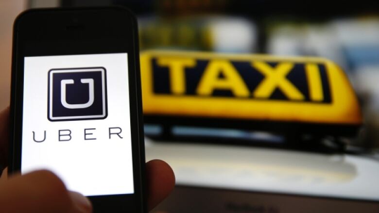 A person holds up a phone with the word 'Uber' on it next to a taxi sign.