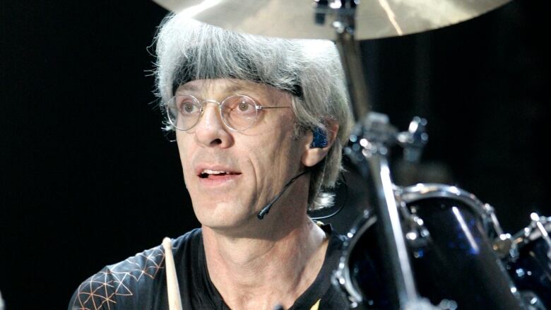 A grey-haired man with glasses is pictured at a drum set.