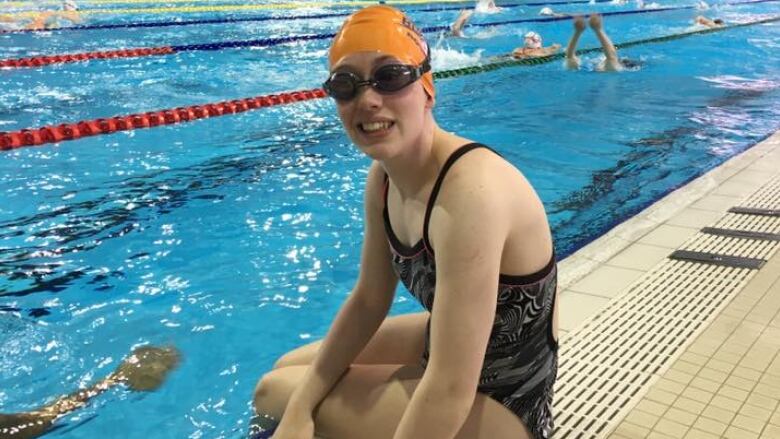 A swimmer wearing an orange bathing cap and goggles sits on the edge of the pool deck