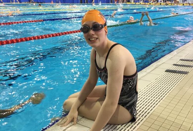 A swimmer wearing an orange bathing cap and goggles sits on the edge of the pool deck