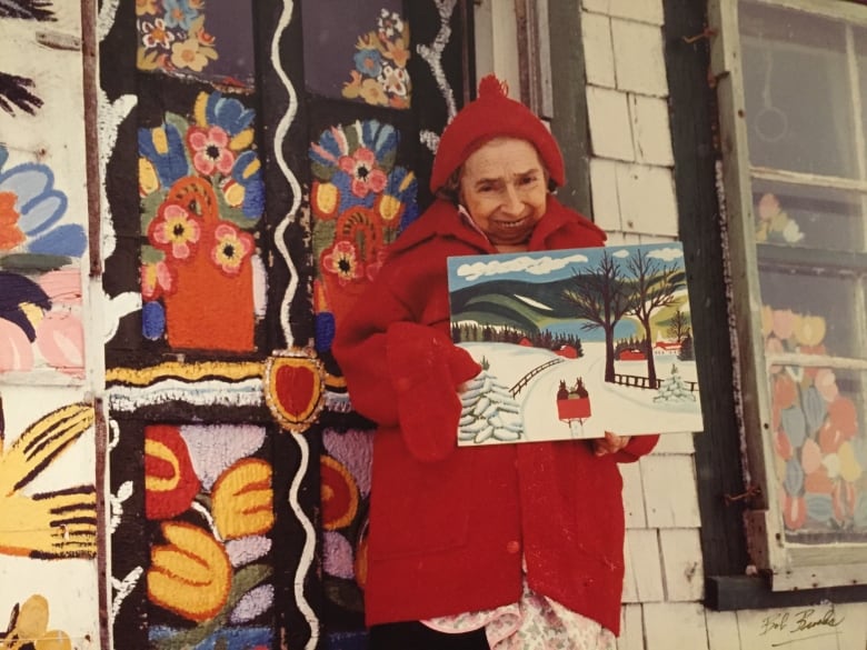 A petite artist is shown with a piece of her art work in front of a colourful house.