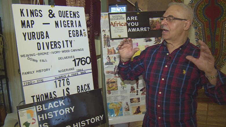 A man with short grey hair and a grey moustache wearing dark glasses and a blue and red plaid button up shirt gestures while speaking about a historical display in front of him.