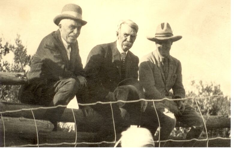 In a black and white photo, three older men sit on a wood fence.