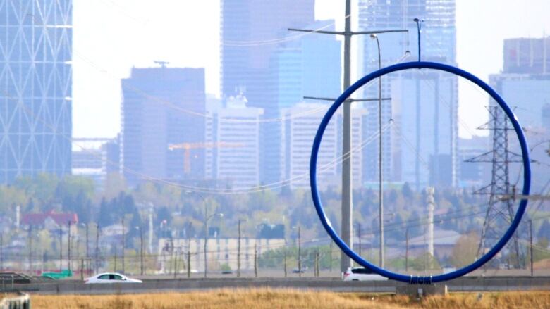 A giant blue ring with what looks like a street lamp on top sits on a road as the Calgary skyline looms in the background.